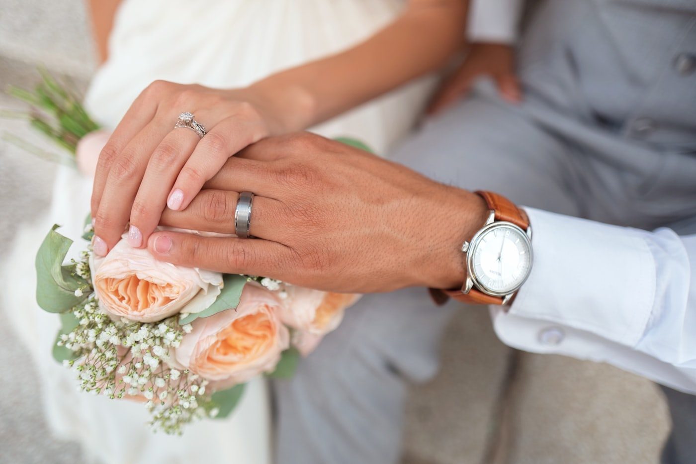 A couple's hands and rings resting on a peach rose bouquet